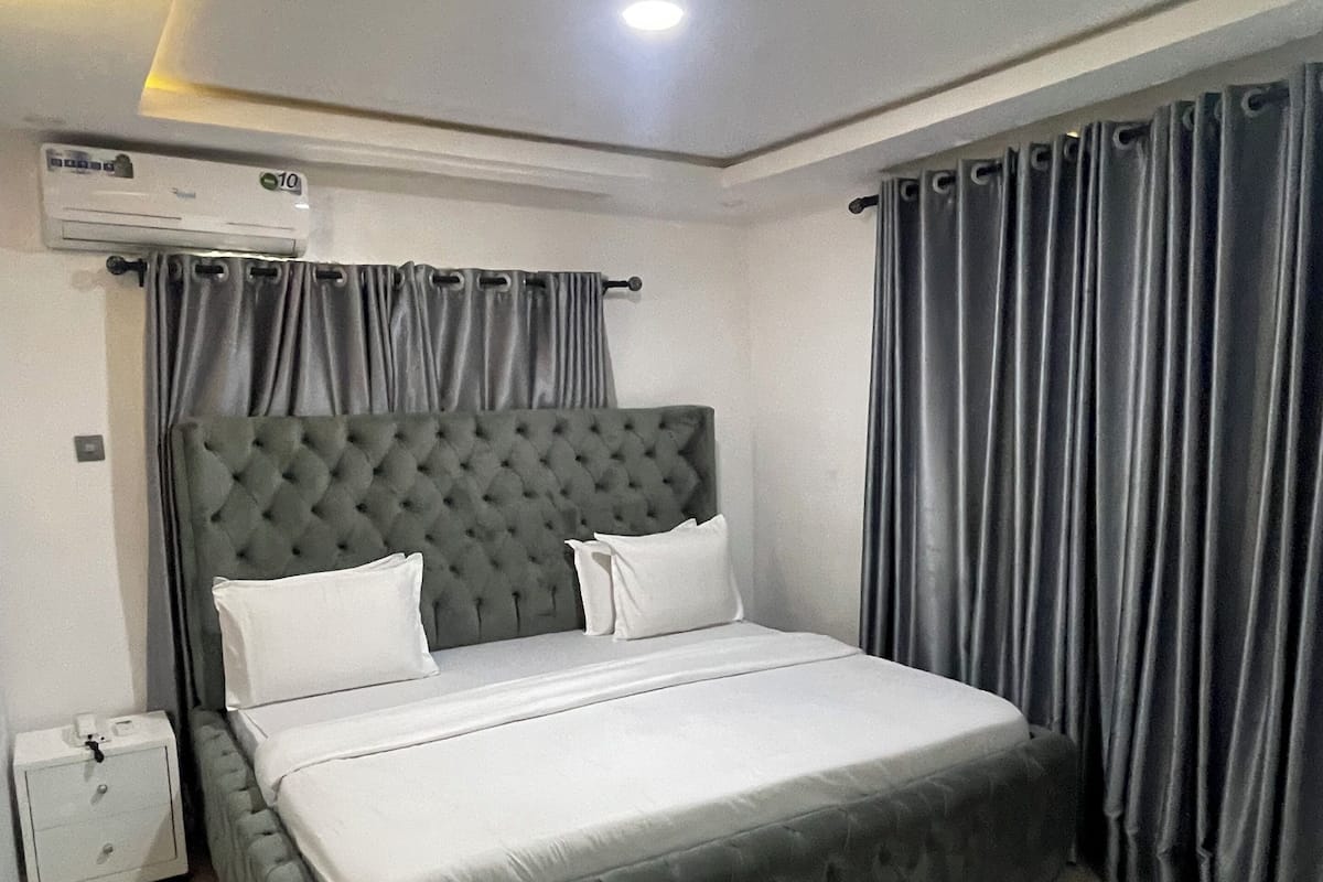 Un dormitorio con una cama grande, cortinas grises, aire acondicionado montado en la pared y una mesita de noche.