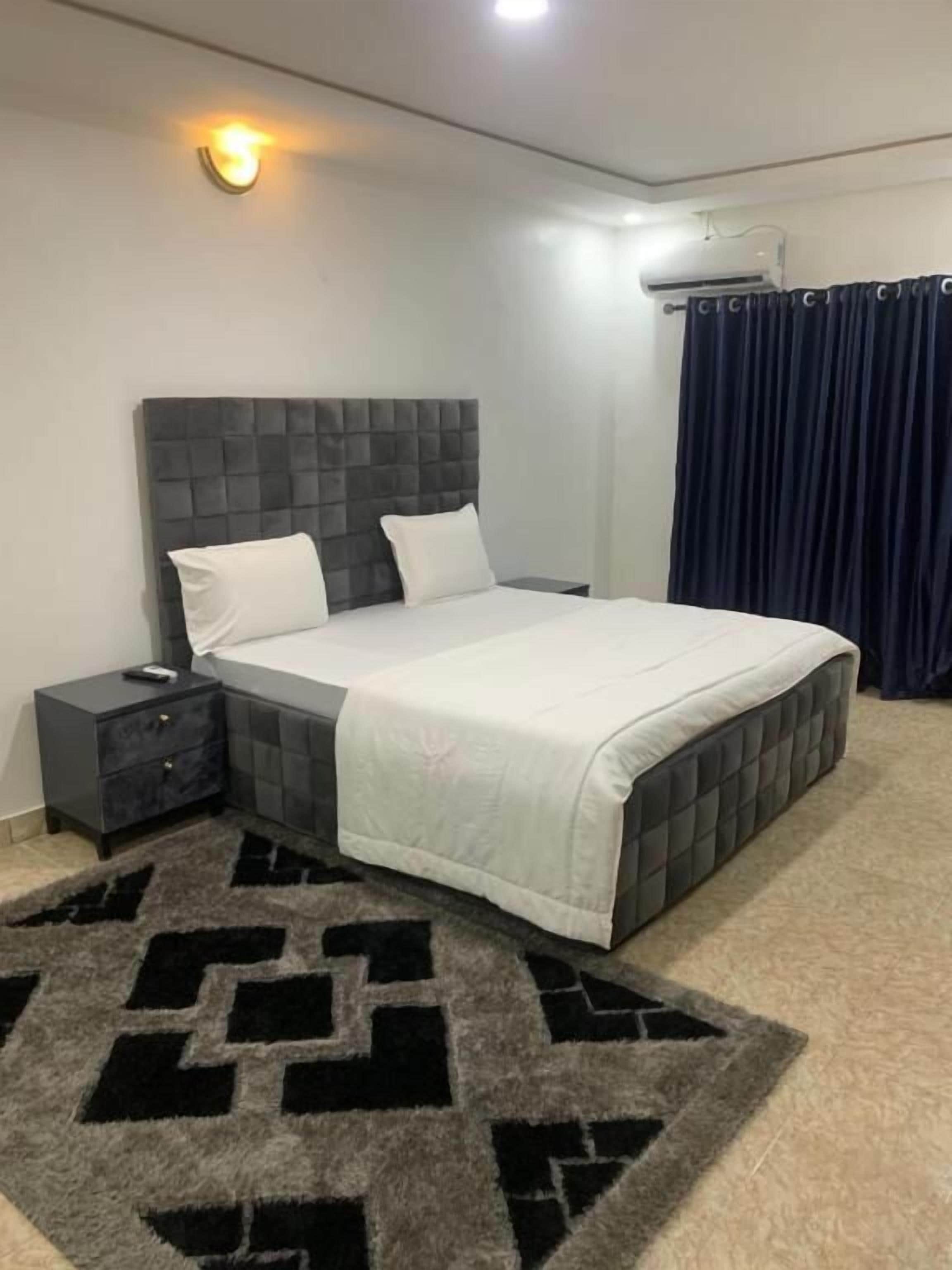 Un dormitorio con cabecera capitonada, cama con ropa de cama blanca, mesita de noche oscura, alfombra estampada y cortina oscura.