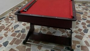 A pool table with a red felt surface, a cue stick, and a set of balls. The room has a tiled floor and a wall with a geometric light fixture. - Weena Hotel & Resort (Lekki)