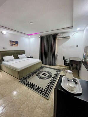 A hotel room with a bed, a desk, a chair, an air conditioning unit, and a coffee maker on a tray. - Weena Hotel & Resort (Lekki)