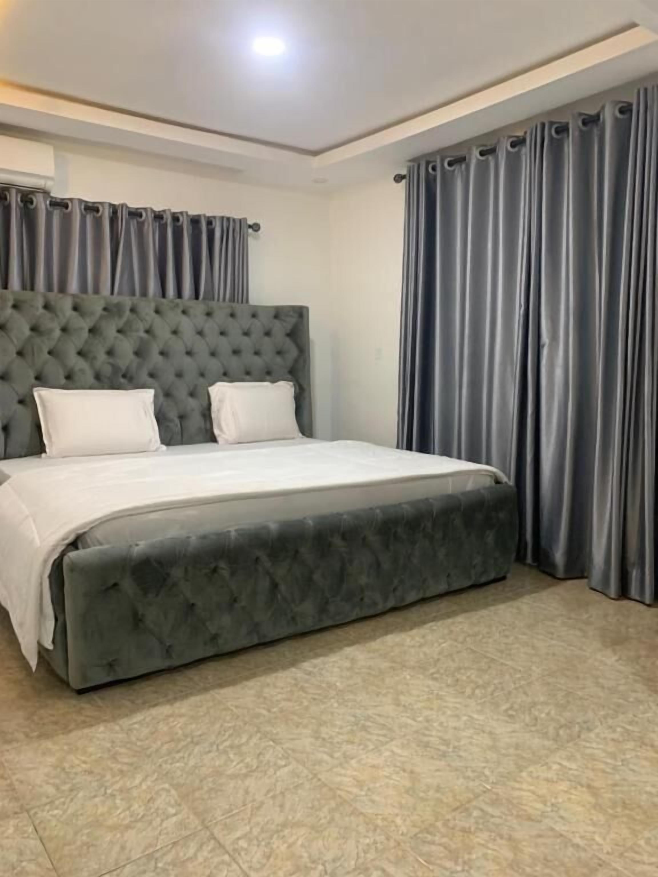 A bedroom with a tufted headboard, a large bed, grey curtains, and a tiled floor.