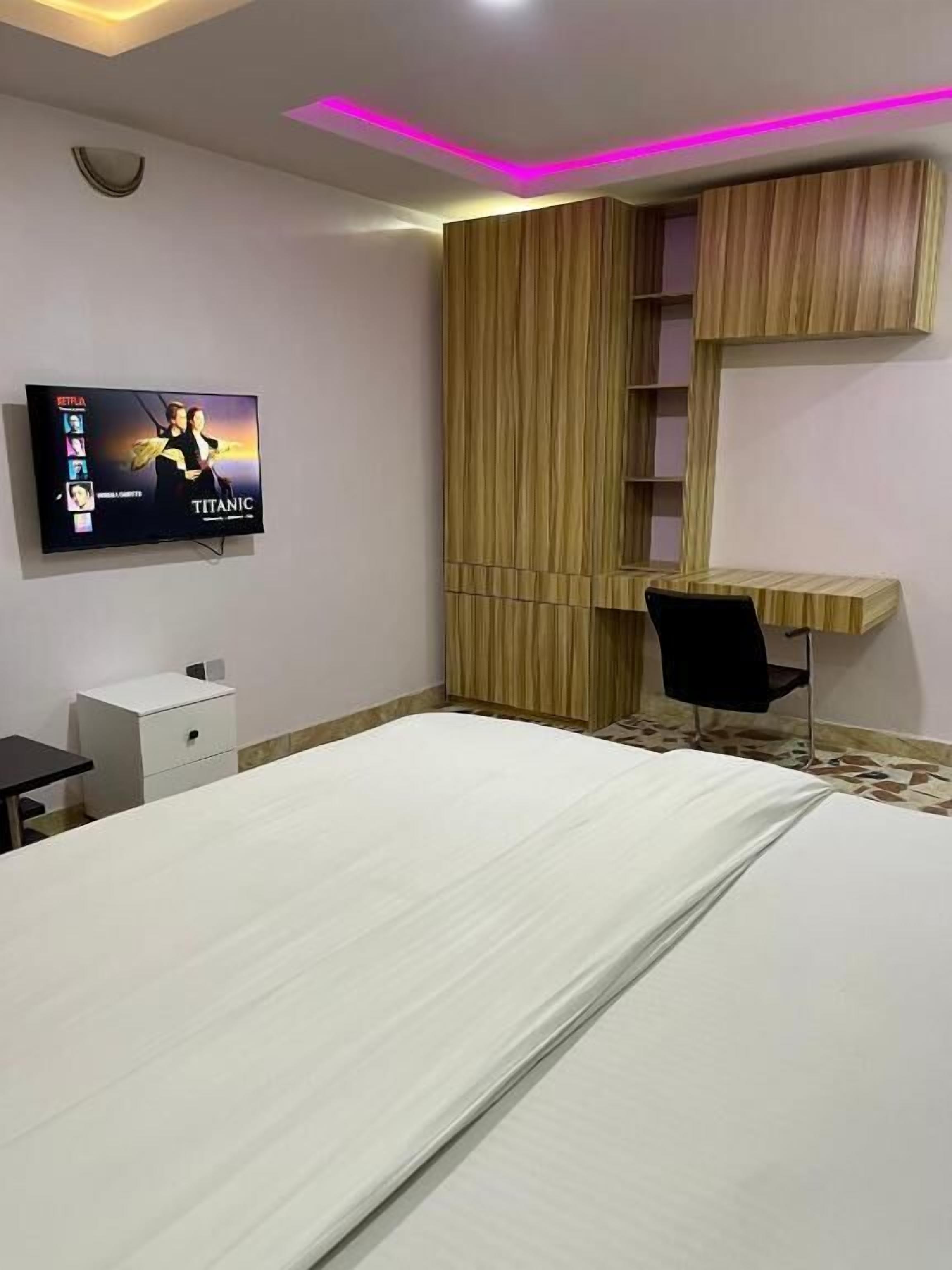 Una habitación de hotel con cama, escritorio, silla, televisor montado en la pared y armario.