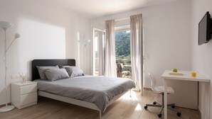 A bedroom with a bed, a desk, a chair, a TV, and a balcony with curtains. - Les Oliviers - 1st Floor Studios by RentalsPro (Vourvourou)