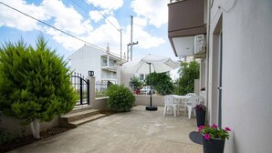 Un patio con una mesa y sillas blancas, un portón negro y una planta en maceta con flores rosadas.