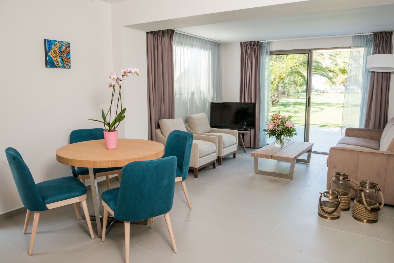 A modern living room with a round wooden table, teal chairs, a beige sofa, a TV, and a balcony with curtains.
