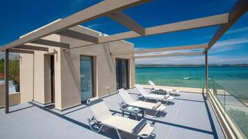 A rooftop terrace with sun loungers, a glass balustrade, and a view of the sea.