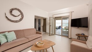 A living room with a sofa, a coffee table, a flat-screen TV, and a view through a glass door to an outdoor area with a chair and a table. - Tiamo Secrets - Beach Front by RentalsPro (Vourvourou)