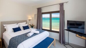 A hotel room with a bed, a TV, a window with curtains, and a view of the sea. - Tiamo Secrets - Beach Front by RentalsPro (Vourvourou)