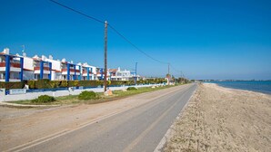 Una carretera costera con una hilera de edificios blancos con detalles azules, una playa de arena y un cielo azul despejado.