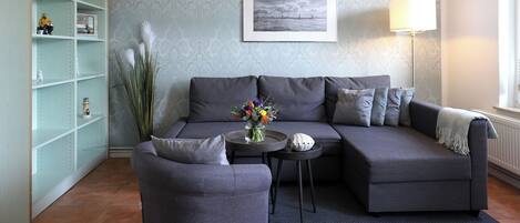 A living room with a grey sofa, a small round table with a vase, a lamp, and a bookshelf.