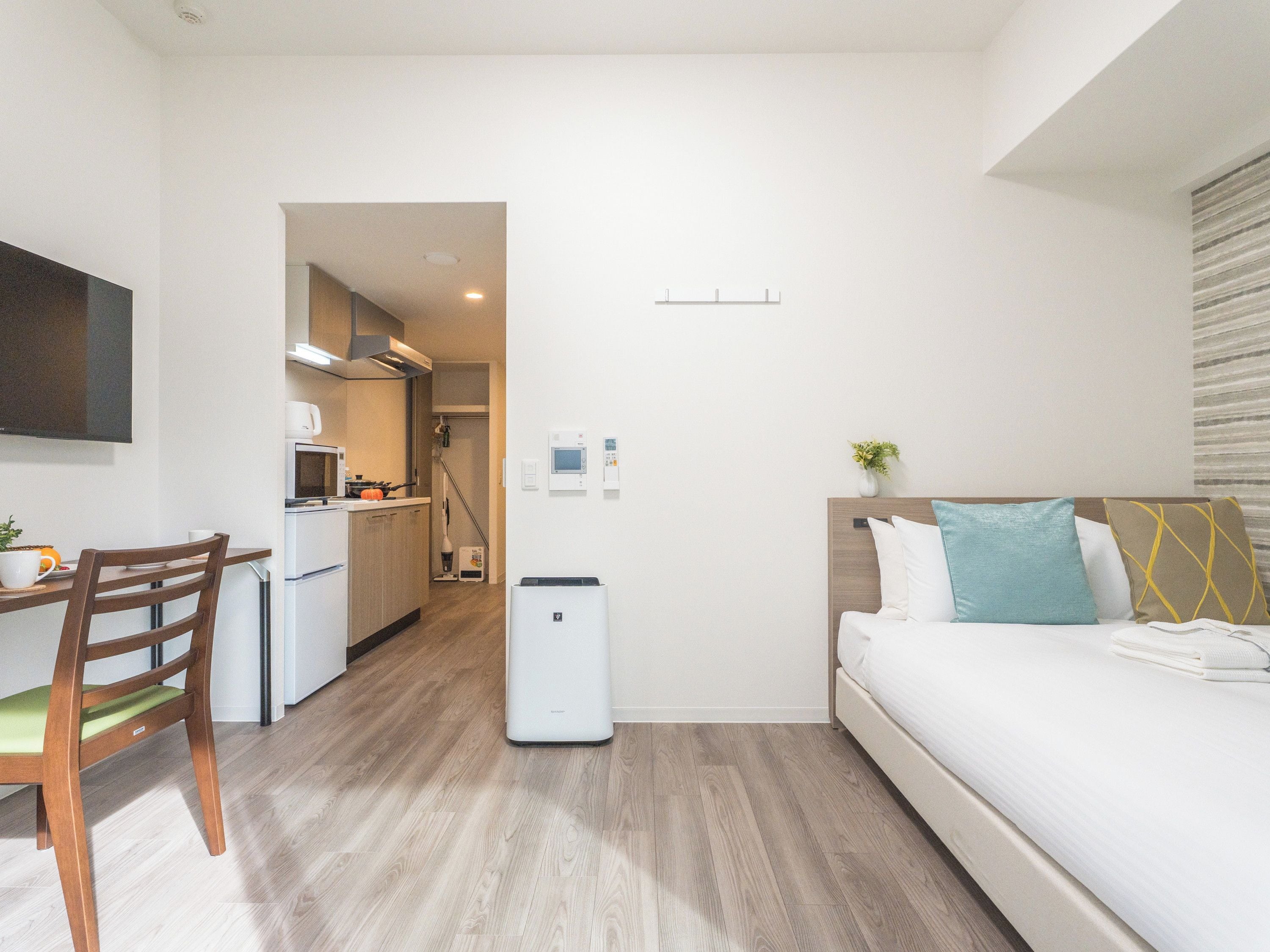 A compact hotel room with a kitchenette, a bed with pillows, and a small table with a chair.
