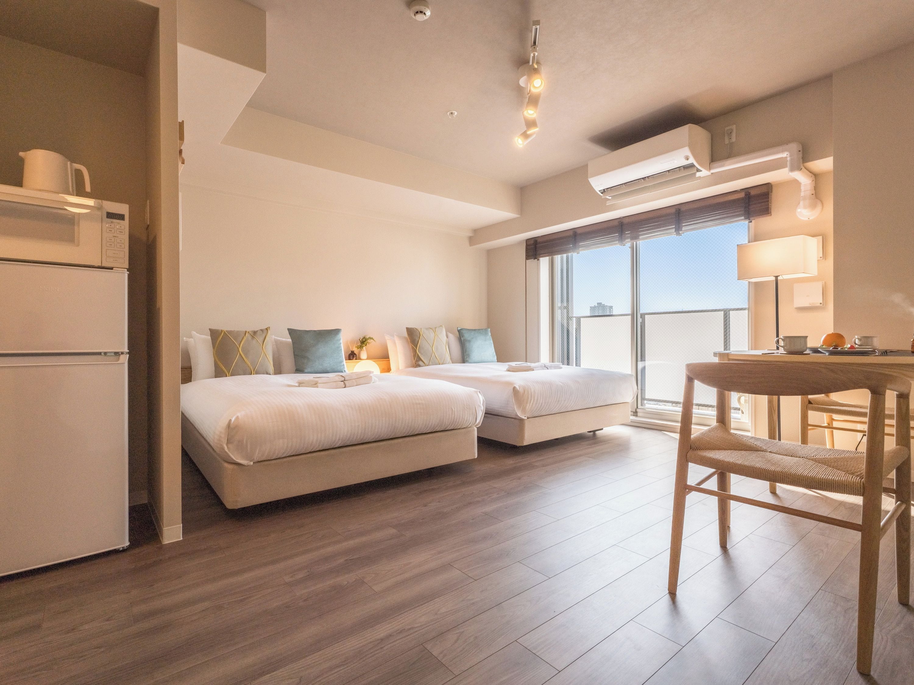 A modern hotel room with a kitchenette, two beds, a dining area, and a large window offering a city view.