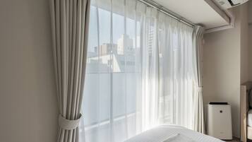 Habitació d'hotel amb un llit, una finestra amb cortines lleugeres, aire condicionat fixat a la paret i una taula de nit amb una làmpada.