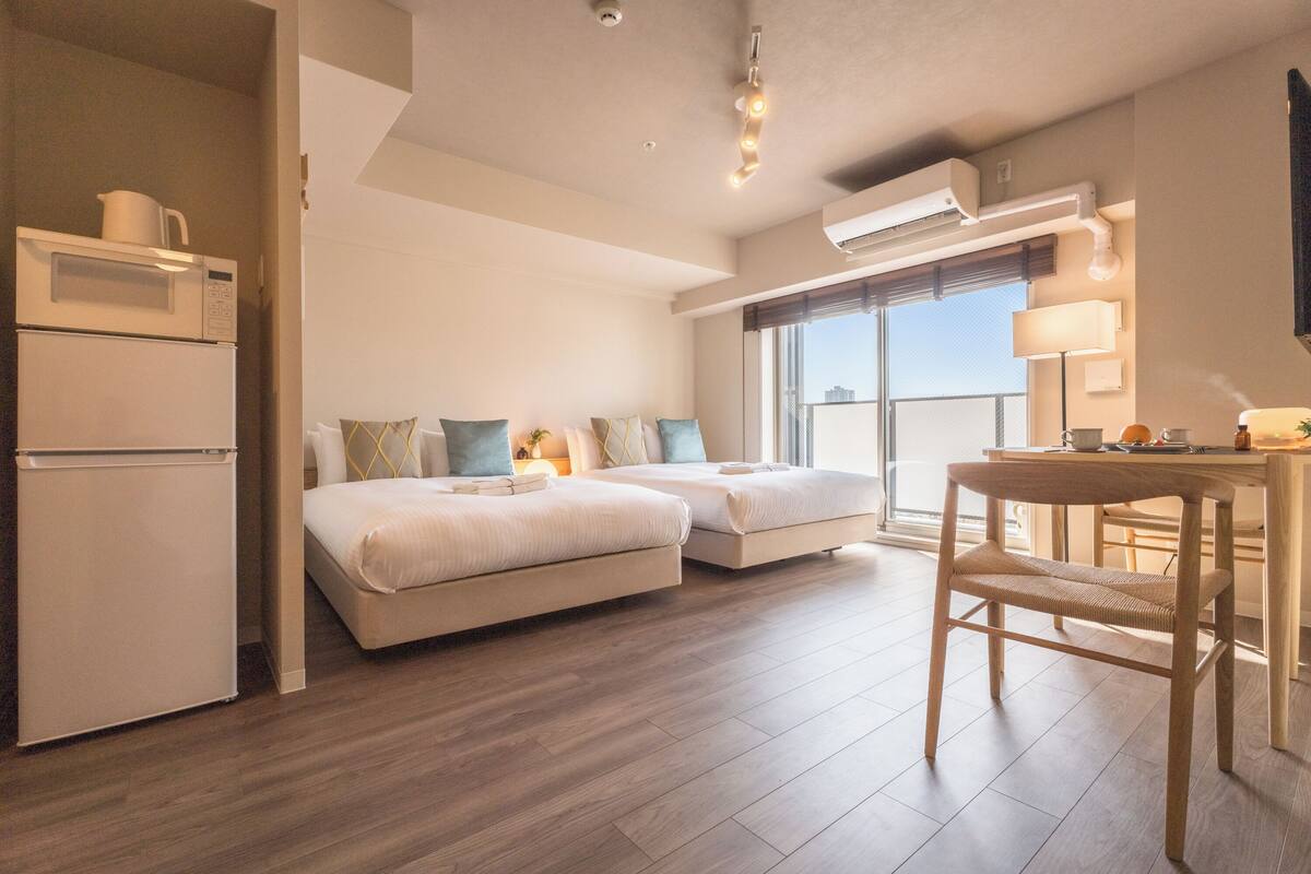 A modern hotel room with a kitchenette, two beds, a dining area, and a large window.