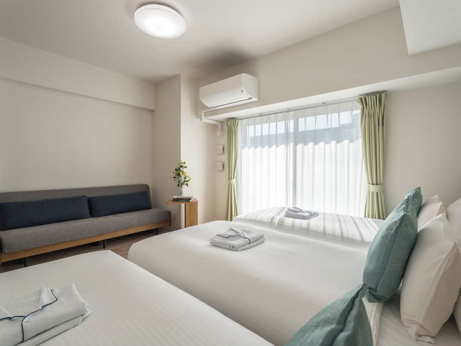 A hotel room with a bed, a sofa, a bedside table with a plant, and an air conditioning unit.