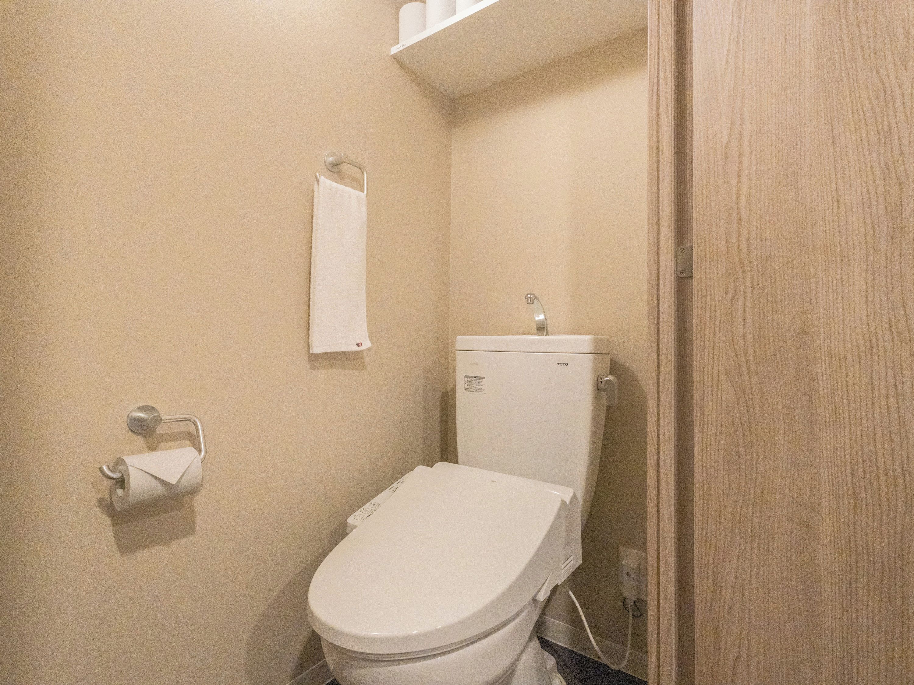A small, compact bathroom with a white toilet, a towel hanging on a metal hook, and a roll of toilet paper on a holder.