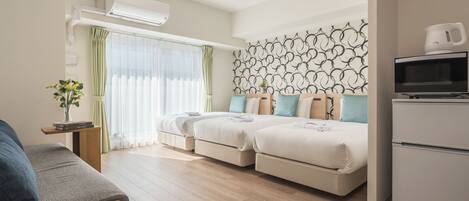 A modern hotel room with a sofa, two beds with matching headboards, a small table with a vase, and a kitchenette.
