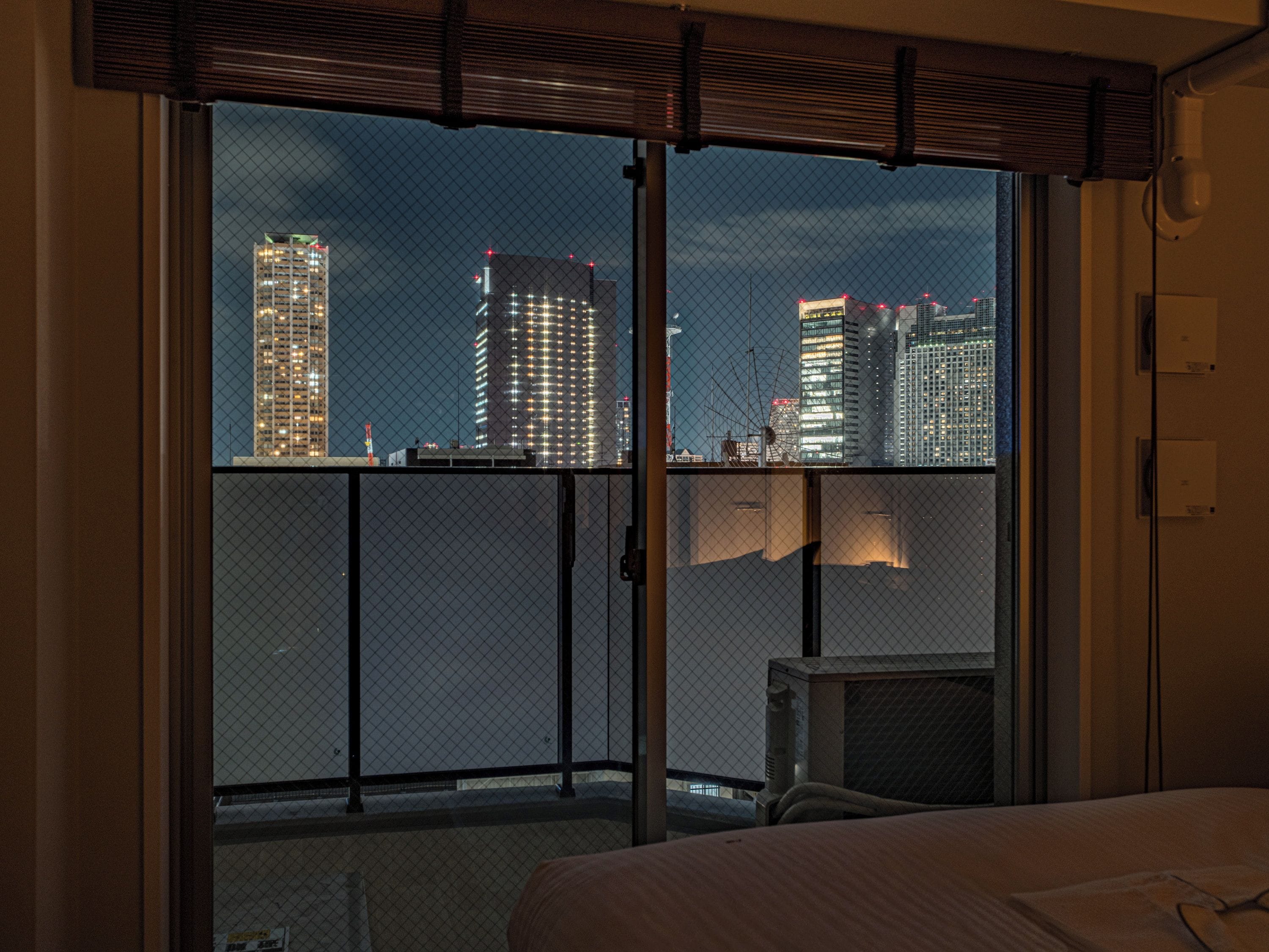 Quarto de hotel com uma grande porta de vidro deslizante que dá para uma varanda com vista para a cidade à noite.