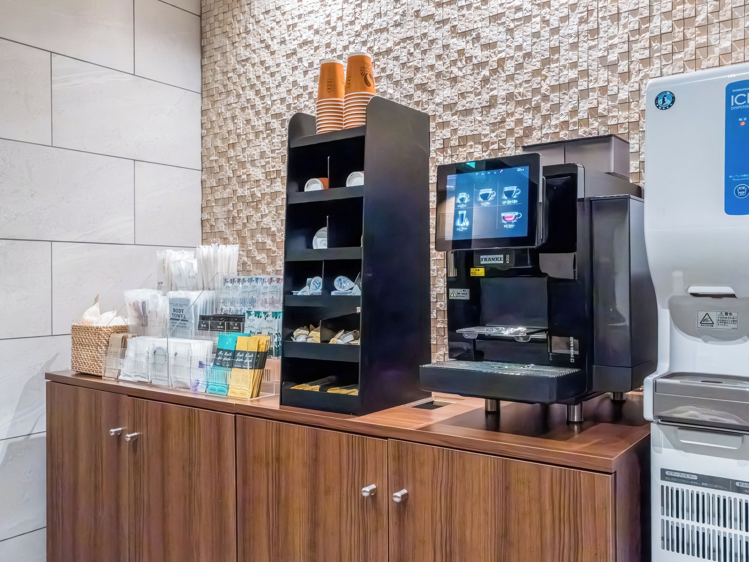 Una estación de café autoservicio con menú digital, dispensador de agua y diversos productos envasados.
