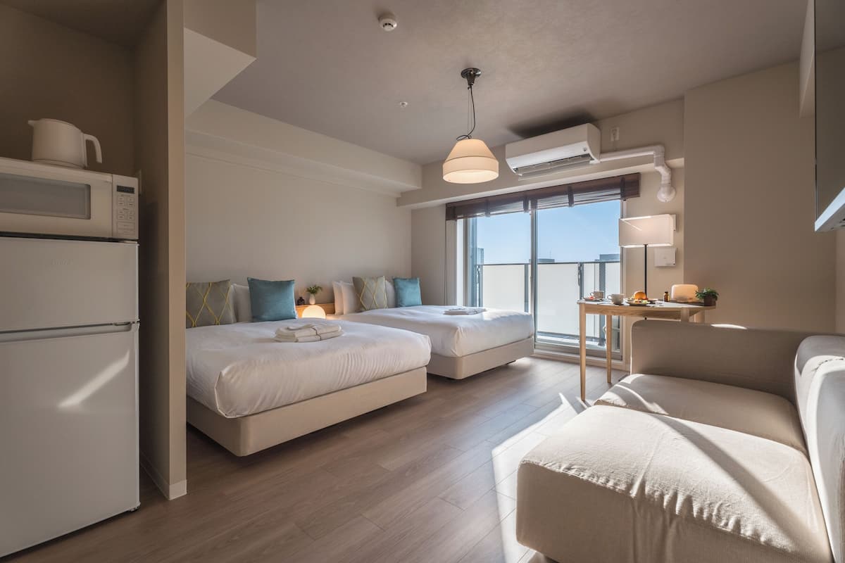 A modern hotel room with a kitchenette, two beds, a sofa, and a balcony with a view.