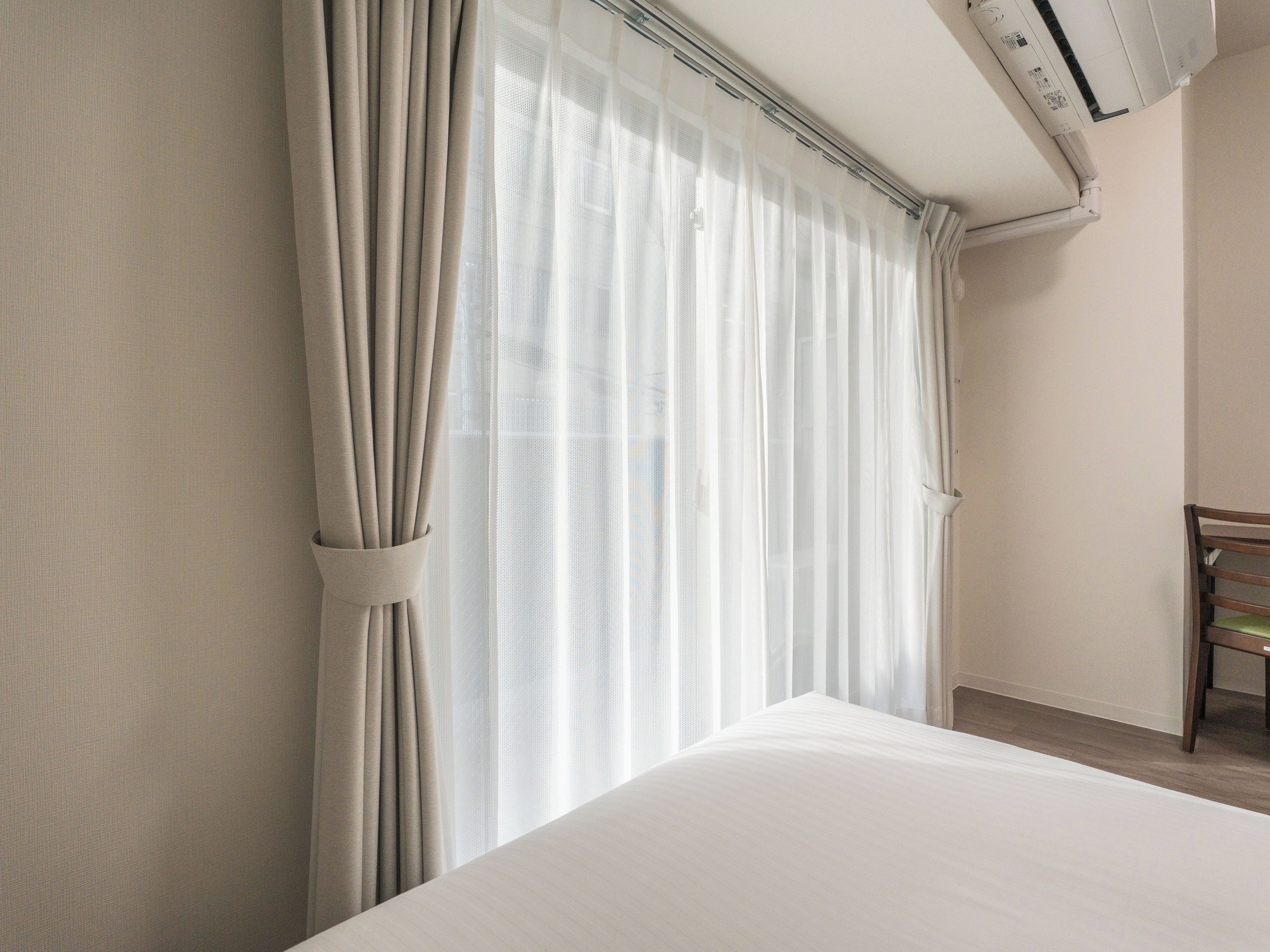 A hotel room with a bed, a chair, and a window with sheer curtains.