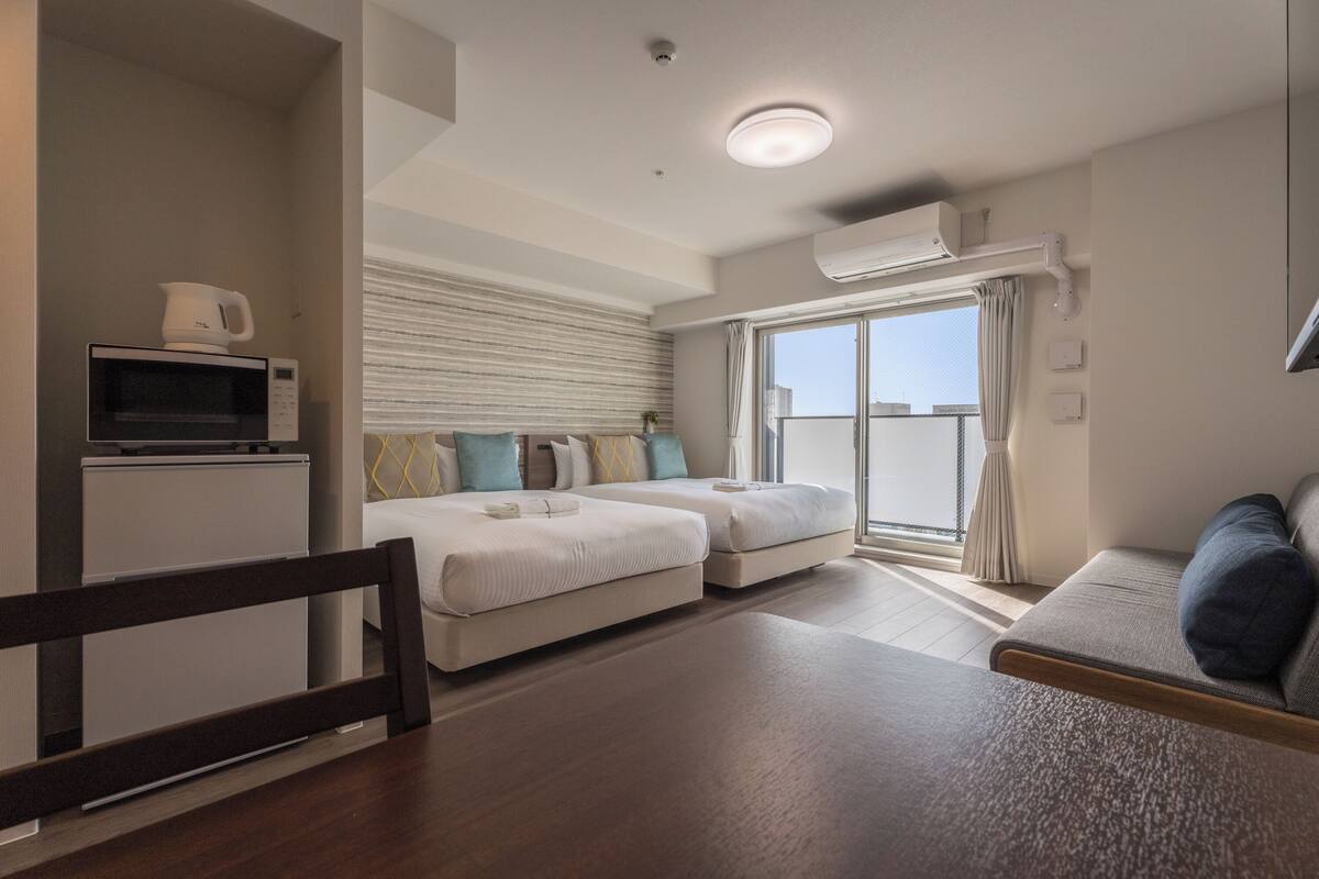 A modern hotel room with a bed, a sofa, a dining table, and a kitchenette.