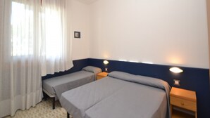 A room with two beds, a window with white curtains, a wall-mounted light, and a small wooden nightstand. - Comfy Flat With Shared Swimming Pool (San Michele al Tagliamento)