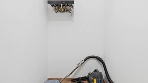Um quarto pequeno com um aquecedor branco fixado na parede, um aspirador de pó e um colchão no chão.