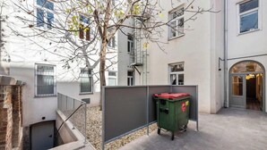 Un patio con un árbol, un bote de basura verde y un edificio con varias ventanas y una puerta.