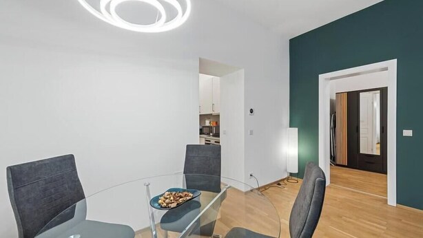 Un comedor moderno con una mesa de vidrio, dos sillas, un tazón de aperitivos, una cocina al fondo y una pared de acento color aguamarina.