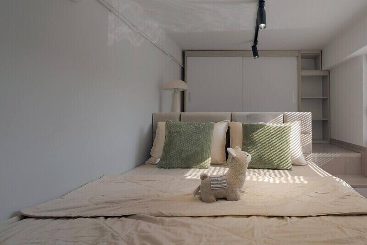 Quarto minimalista com cama, almofadas, um pequeno brinquedo de pelúcia e uma luminária de cabeceira.