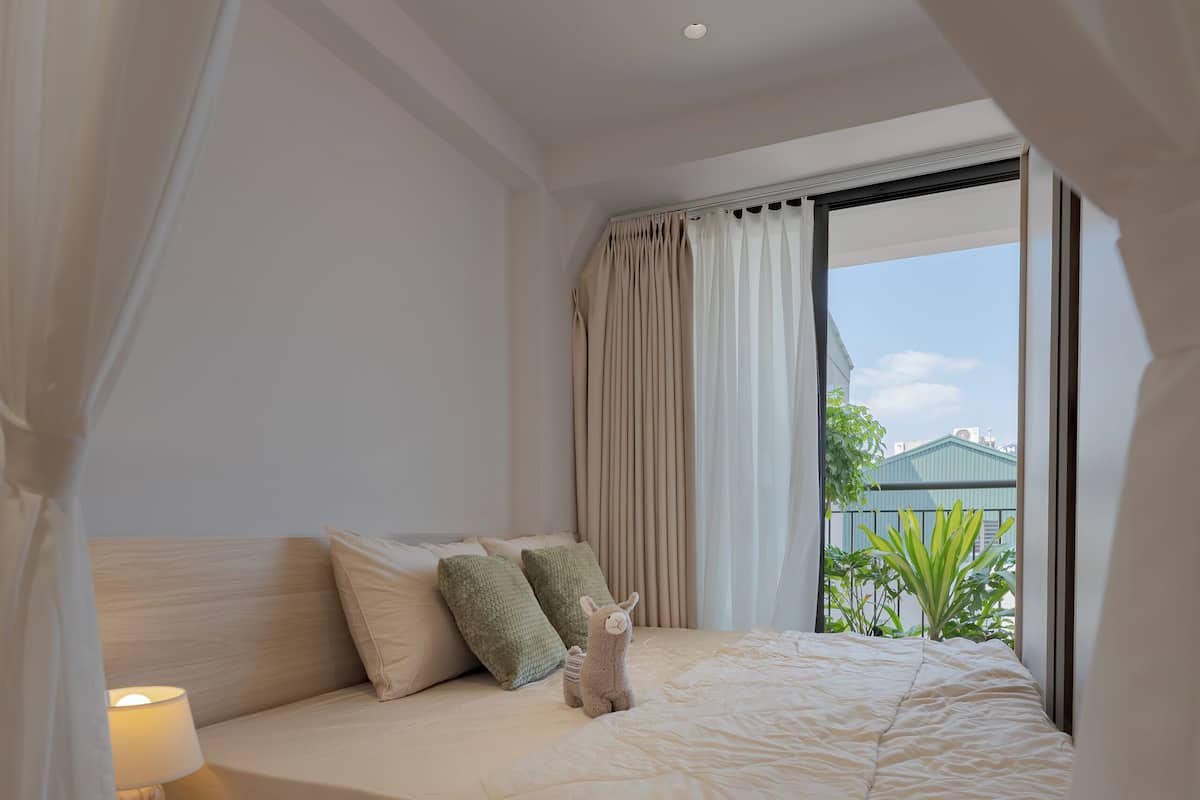 Quarto com cama, cortinas, luminária e vista para o verde lá fora.