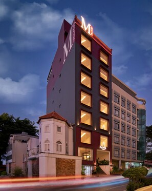 A multi-story hotel with a prominent 'M' sign, multiple illuminated windows, and a modern architectural design. - Express by M Village Hoàng Việt, a brand of Modern Village Lifestyle (Ho Chi Minh)