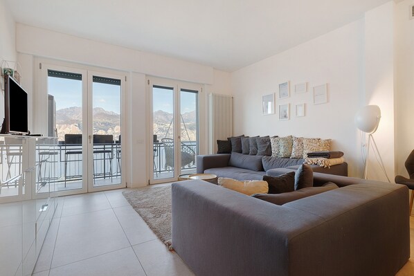 Ein modernes Wohnzimmer mit grauem Sofa, einem Flachbildfernseher und einem Balkon mit Blick auf die Berge.