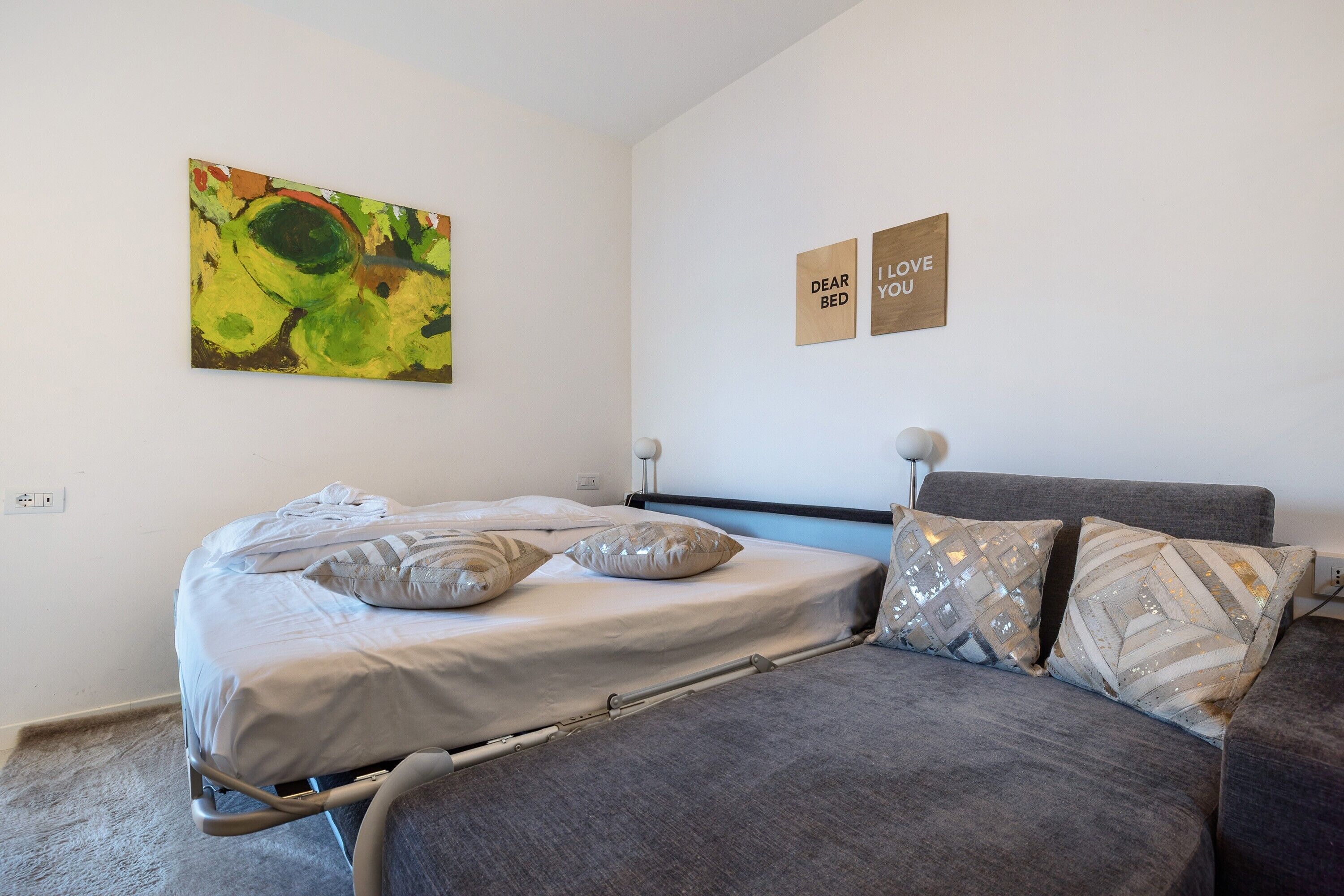 Un dormitorio con cama, dos almohadas, una mesita de noche, una lámpara y un cuadro en la pared.
