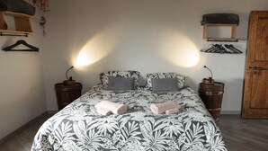 Cama com colcha estampada, duas mesas de cabeceira com abajures, guarda-roupa de madeira e prateleira de madeira com uma peça de roupa pendurada.