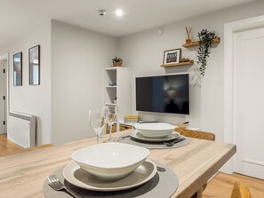 A modern dining area with a wooden table set for two, a television mounted on the wall, and a white radiator in the background. - Village Centre Haven (Clifden)