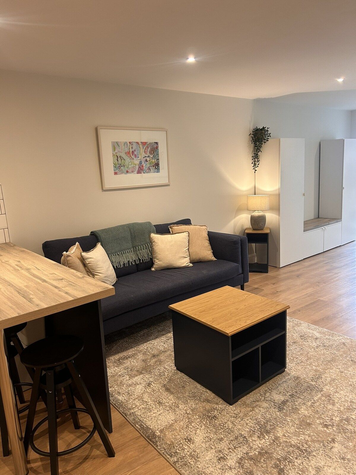 A modern living room with a sofa, a coffee table, a dining table with chairs, a framed artwork, a floor lamp, and a mirror.