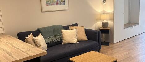 A modern living room with a sofa, a coffee table, a dining table with chairs, a framed artwork, a floor lamp, and a mirror.