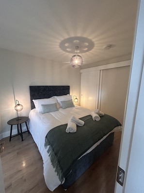 A neatly made bed with pillows and a blanket, a bedside table with a lamp, and a ceiling light fixture. - Village Centre Haven One Bed Apartment (Clifden)