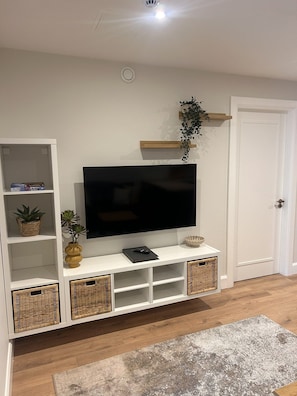 Ein Wohnzimmer mit einem Flachbildfernseher auf einer weißen Unterhaltungseinheit, Weidenkörben, einer Pflanze und einer Tür.