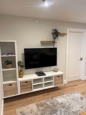 A living room with a flat-screen TV, wicker baskets, a potted plant, and a door. - Village Centre Haven One Bed Apartment (Clifden)