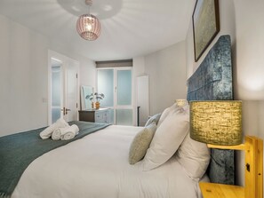 A bedroom with a bed, a bedside lamp, a dresser, and a view of a bathroom. - Village Centre Haven (Clifden)
