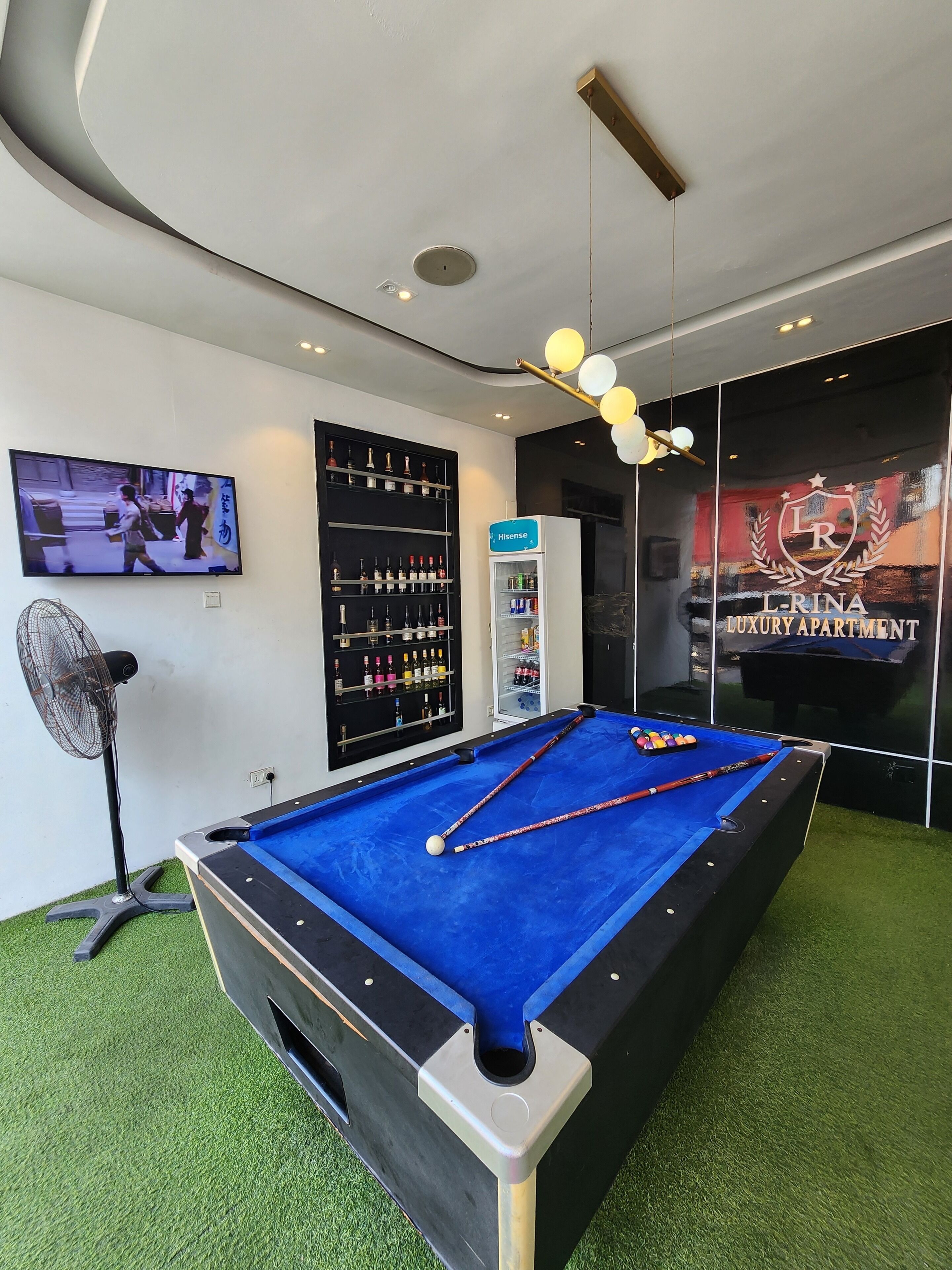 A pool table with cues and balls, a television, a fan, a minibar, and a wall with a liquor display.