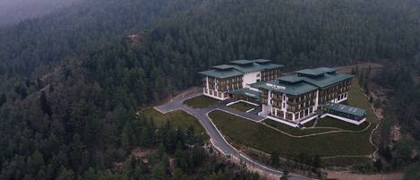A large, modern building with multiple sections, surrounded by dense forest and hills.