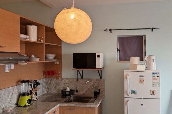 A compact kitchen with a microwave, refrigerator, and a hanging light fixture. - SeafrontView · Cozy T1 Apartment - 1 (Praia)