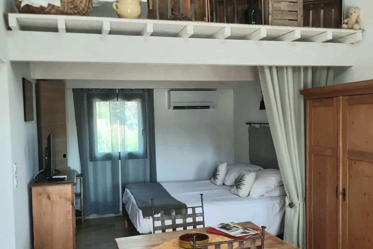 A dorm-style room with a loft bed, a desk, a chair, a small table, and a wardrobe.