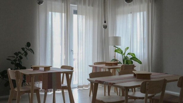 Un espace repas avec des tables et des chaises en bois, de grandes fenêtres ornées de rideaux légers et des lampes suspendues.
