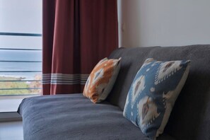 A grey sofa with two patterned cushions, a window with a view, and a red curtain. - SeafrontView · Cozy T1 Apartment - 2 (Praia)