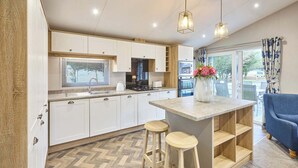 A modern kitchen with white cabinets, a central island with stools, a built-in oven, and a window with blinds. - Host & Stay | The Haven Lodge (Pocklington)